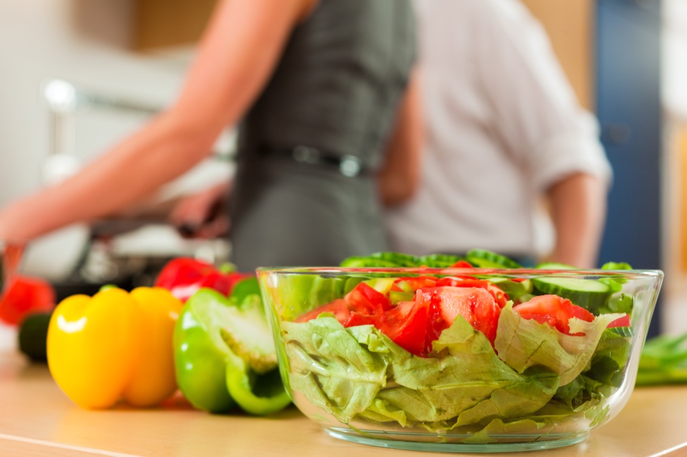 Frischer Salat in einer Glasschüssel, im Hintergrund kochen zwei Personen